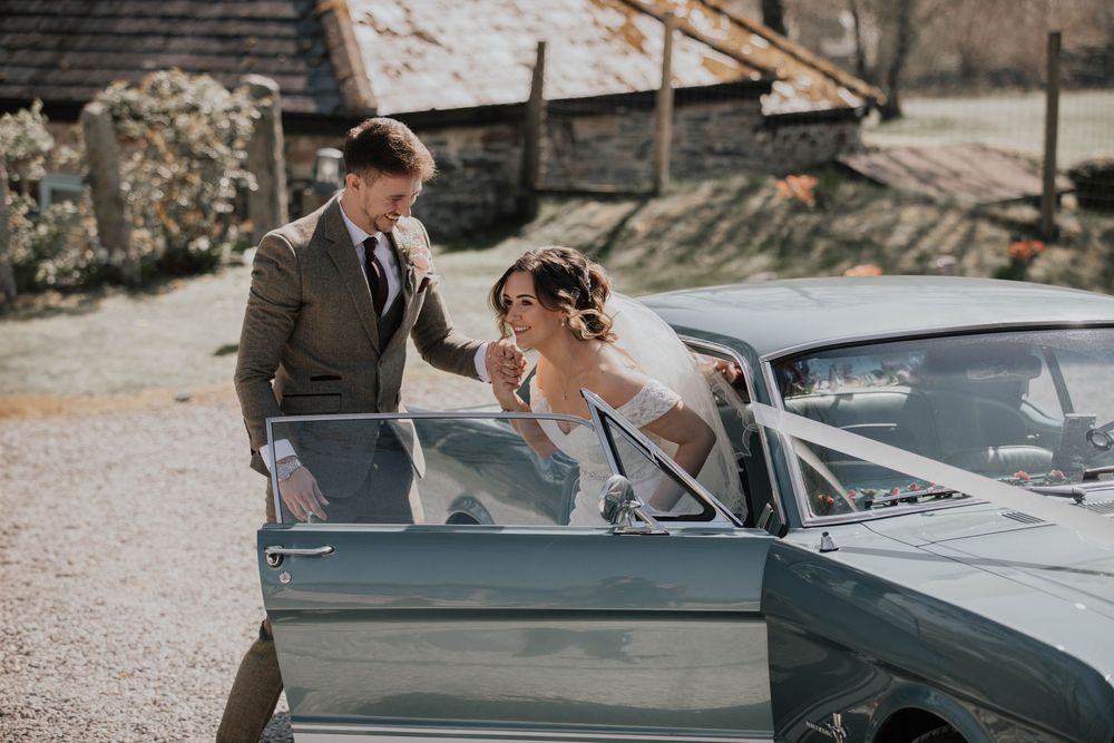 Groom helps bride in white wedding dress exit a vintage teal convertible car on wedding day.