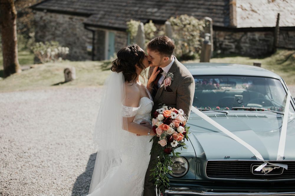 Bride and groom share a kiss beside a classic green Ford Mustang decorated with white ribbon.