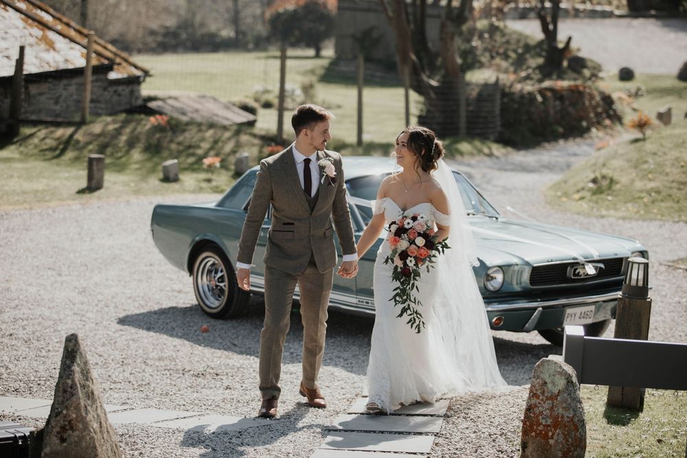 Bride and groom hold hands walking past a vintage green Ford Mustang at rustic wedding venue.