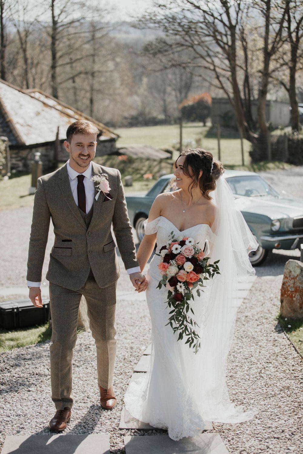 Smiling bride and groom walk hand in hand at rustic outdoor wedding venue on sunny spring day.