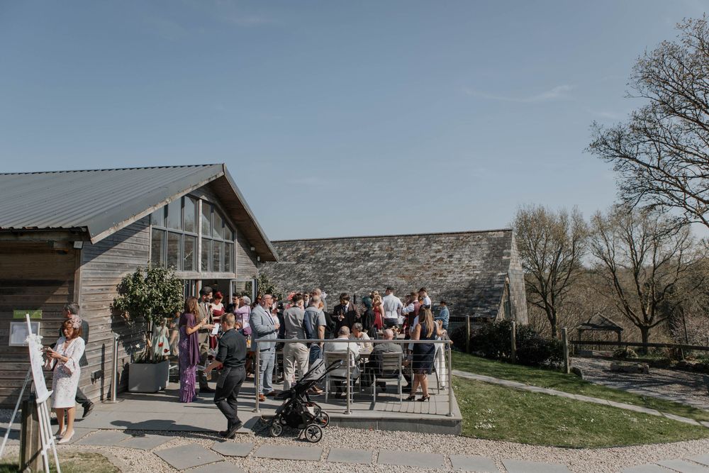 Wedding guests mingle outside a modern rustic barn venue with stone walls on a sunny day.