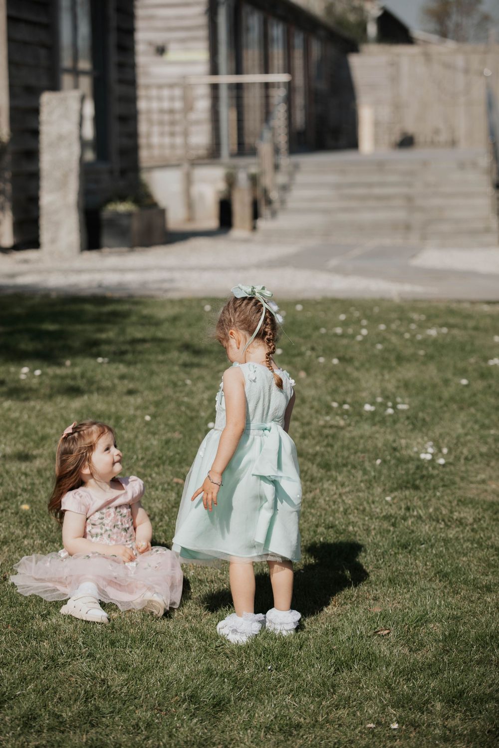Two young flower girls in pastel dresses playing on green grass at an outdoor wedding venue.