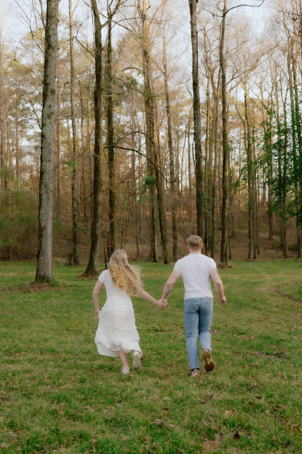 Playful couple running and dancing through a sunlit forest clearing with tall bare trees in the background.