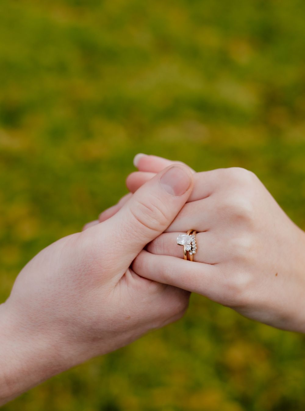 Two hands gently touch while displaying an elegant engagement ring against a soft green grass background.