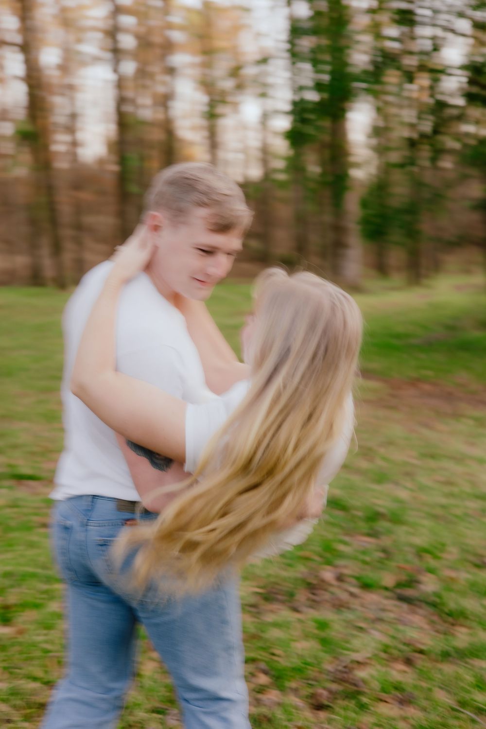 A couple dancing and spinning together playfully in a wooded outdoor setting wearing casual denim and white clothing.