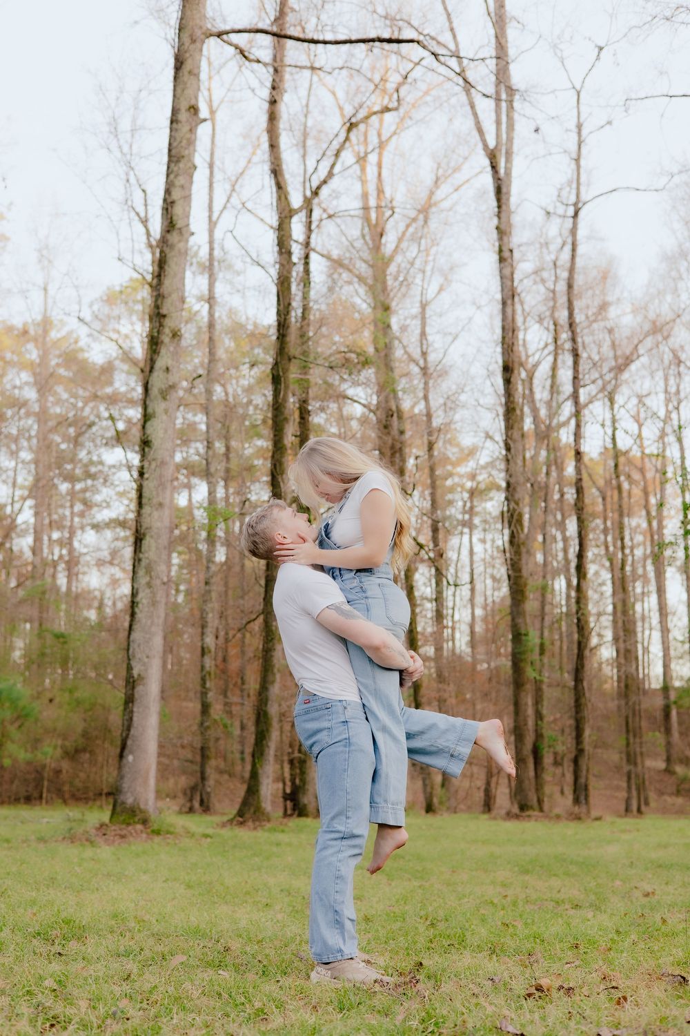 A couple shares a romantic embrace in a wooded area with bare trees and green grass in early spring.