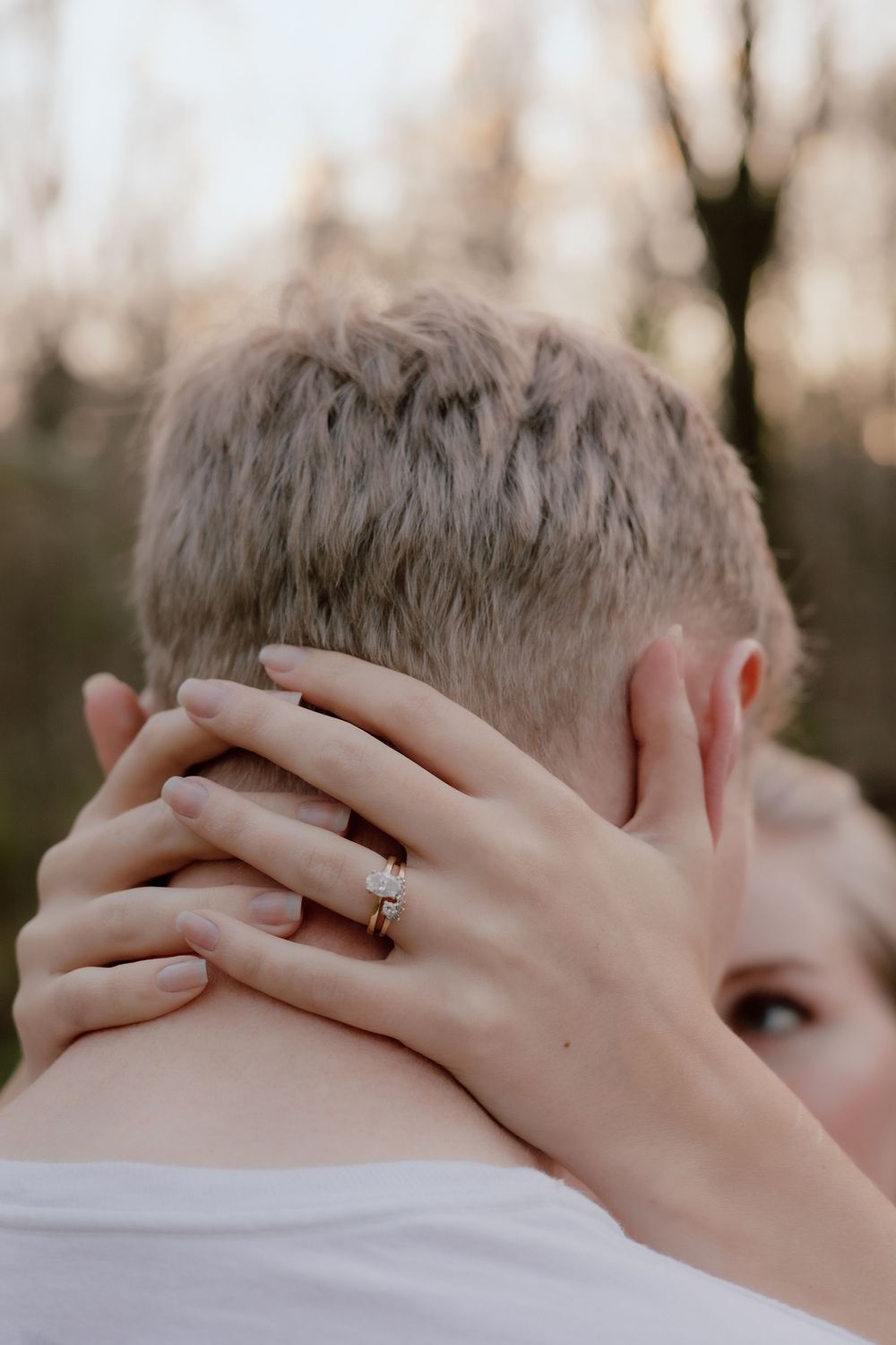 A couple shares a tender kiss while showing off their engagement rings in a romantic outdoor setting at sunset.
