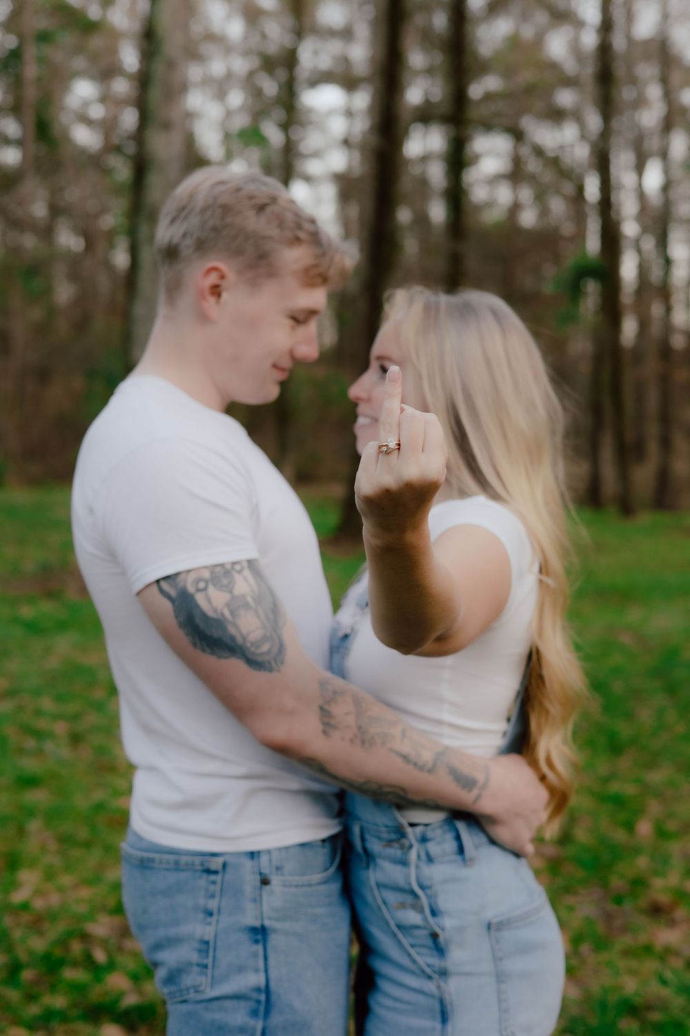 A couple in matching white shirts and jeans embrace in a wooded outdoor setting surrounded by trees.