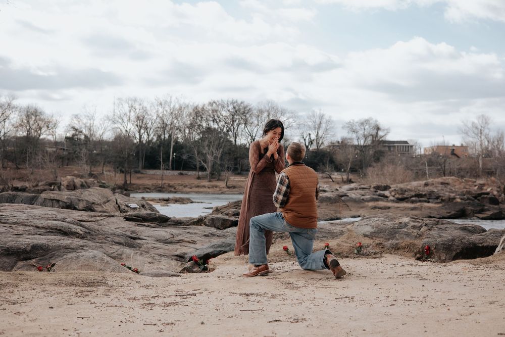 A photographer crouches on a sandy beach with camera equipment during an overcast day with bare trees in background.