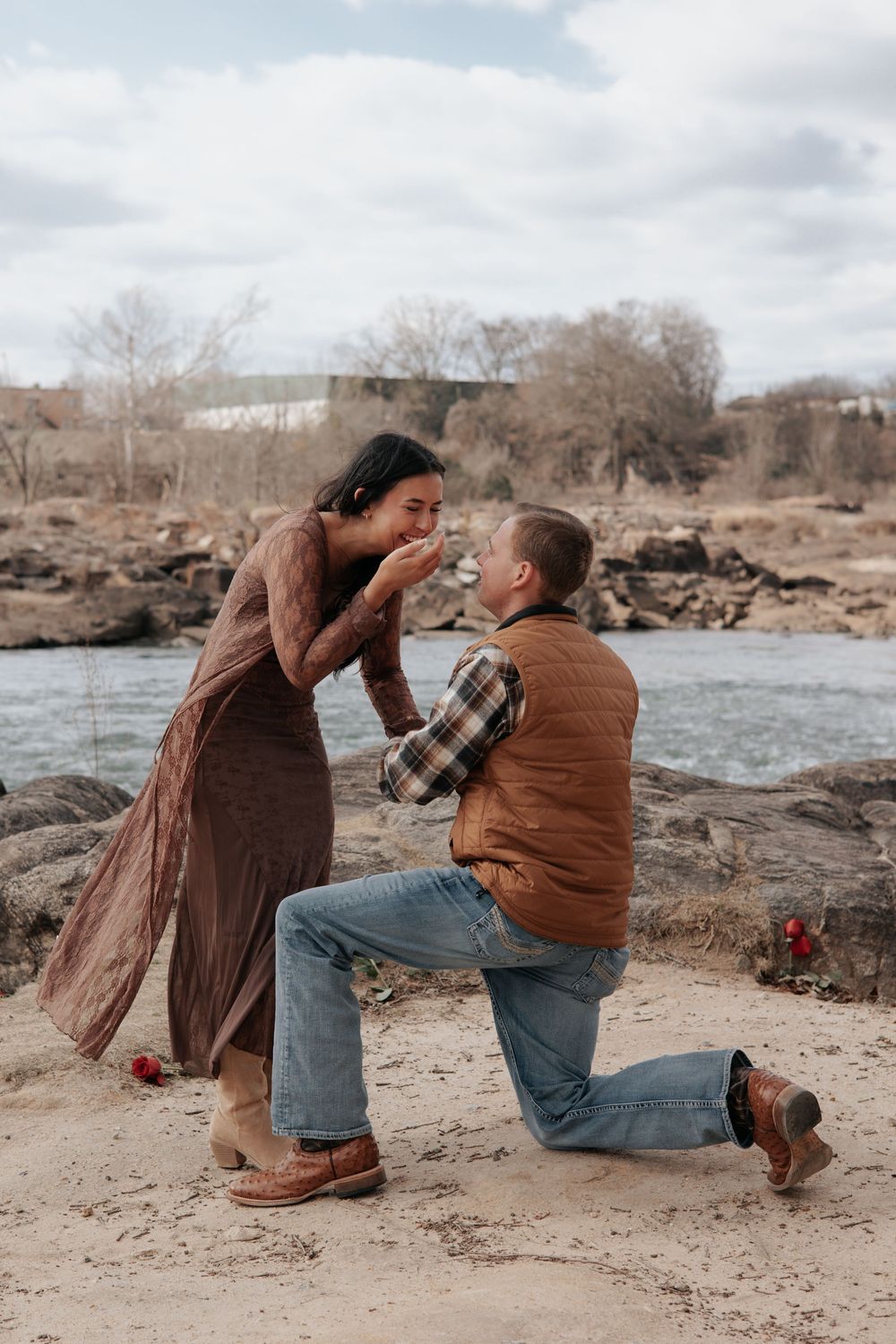 A couple shares a romantic moment during a proposal by a rocky riverbank on an autumn day.