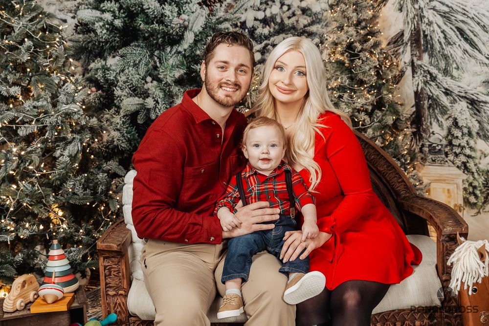 Family wearing red and beige outfits poses for holiday photos in front of decorated Christmas trees and winter decor. Holiday Photos Webster