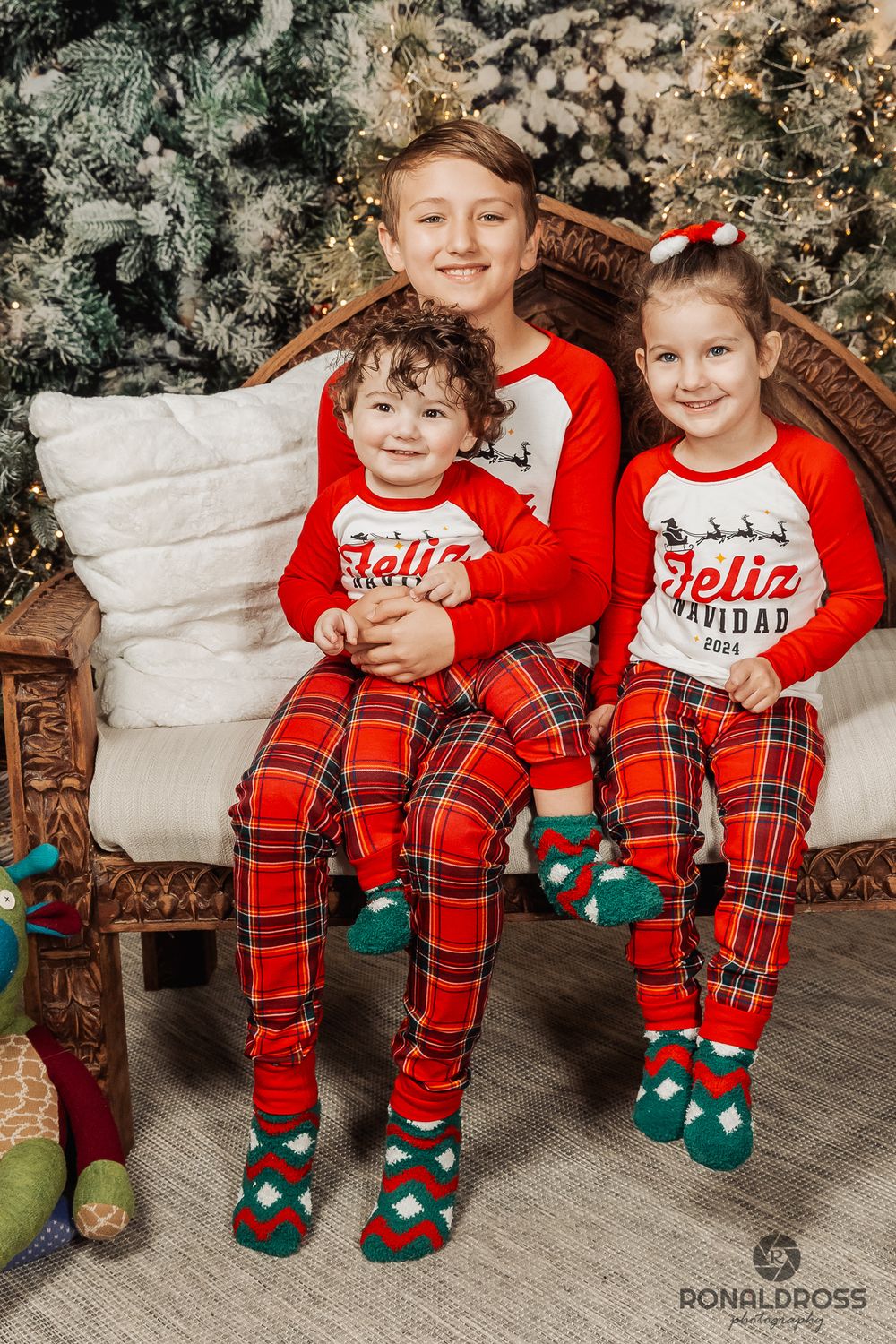 Two children wearing matching red and plaid Christmas pajamas and elf socks sit together by a decorated tree. Holiday Photos Webster