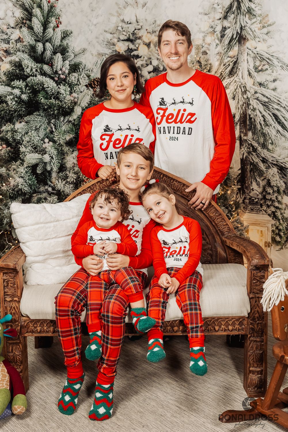 Family wearing matching red and white Feliz Christmas shirts and plaid pajama pants pose on wooden bench with winter backdrop. Holiday Photo
