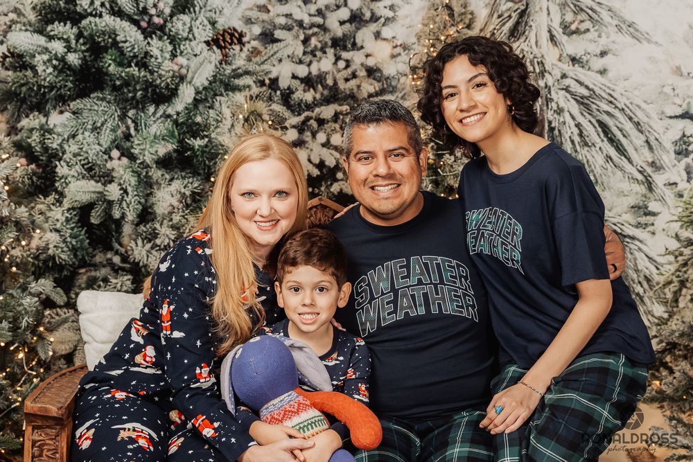 A family wearing coordinated blue and black outfits poses together outdoors against a stone wall background. Christmas Photos Webster