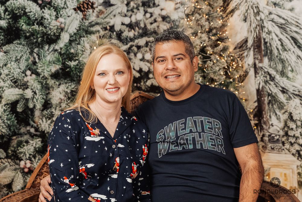 A couple poses together in front of snow-covered evergreen trees during a winter photoshoot. Holiday Photos League City, Webster, Galveston