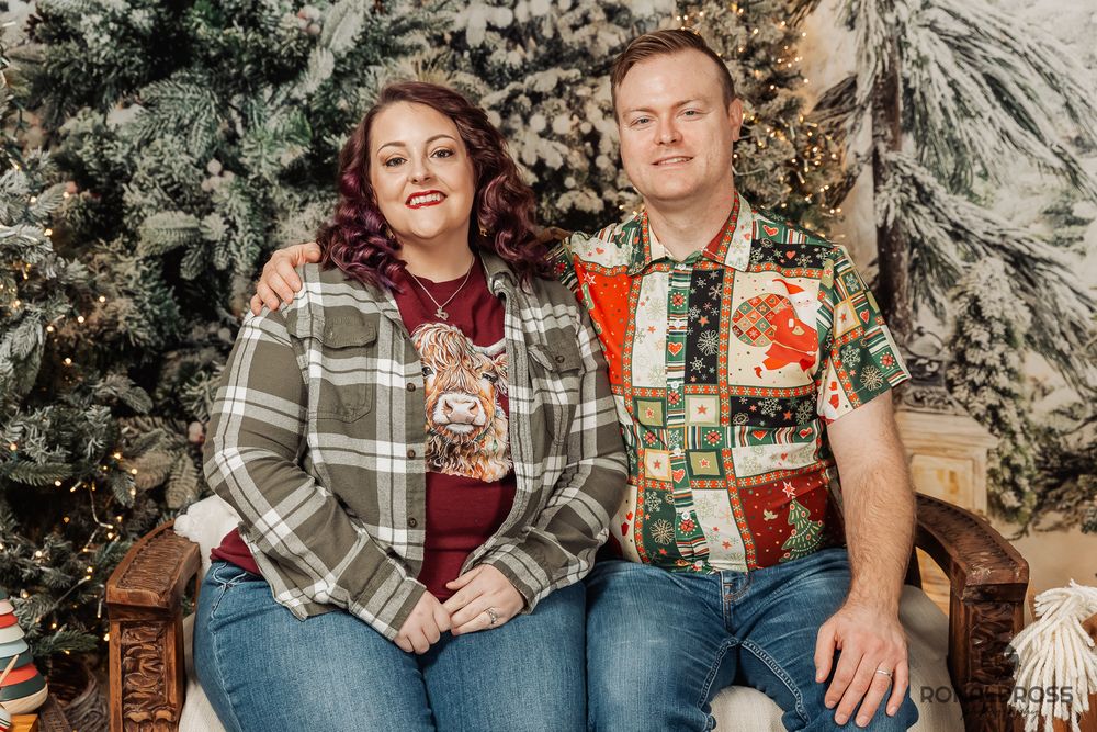 A couple in plaid and flannel shirts sits together on a rustic wooden bench against a snowy winter backdrop. Holiday Photos League City
