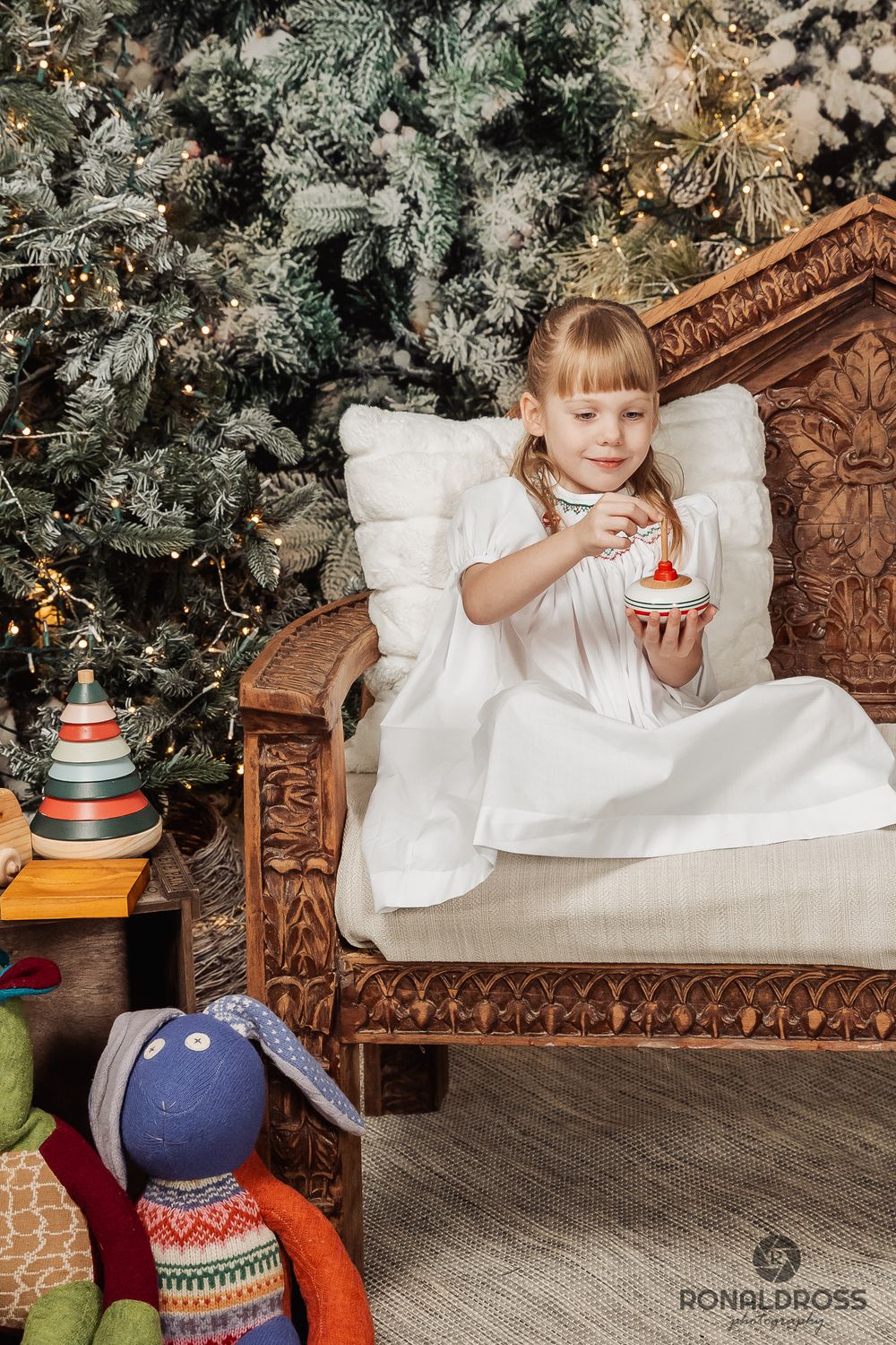 Festive holiday scene with child in white dress sitting on ornate chair next to decorated Christmas tree and toys. Christmas Picture Webster