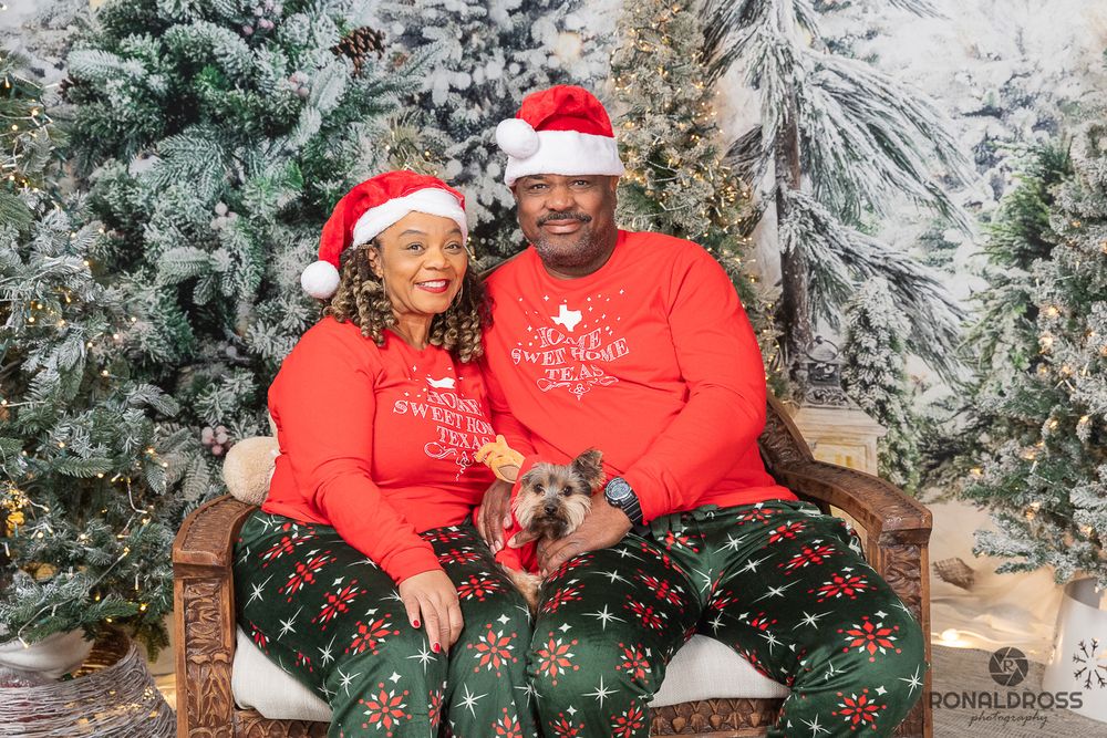A couple wearing matching red sweaters and festive pajama pants pose on a bench amid snow-covered Christmas trees. Holiday Photo League City