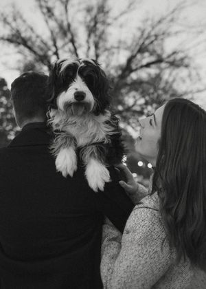 A couple holds a small white and gray dog while standing together outdoors in a casual setting.