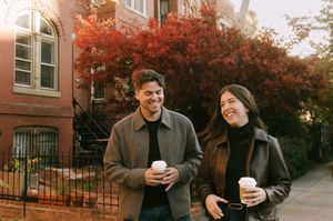 People walking together and laughing while holding coffee cups on an autumn street with red-leaved trees.