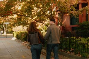 A couple walks along a tree-lined sidewalk on an autumn afternoon with golden sunlight filtering through the leaves.