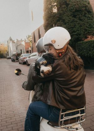 Person in brown jacket and white helmet sitting on a parked scooter on brick pavement.