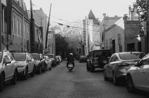 Black and white urban street scene with parked cars and buildings lining both sides of the road.