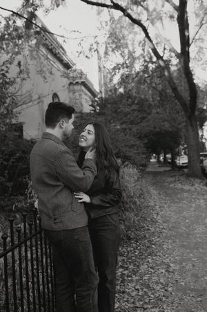 A romantic moment captured in black and white near an iron fence surrounded by autumn trees.