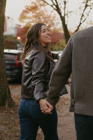 A couple embraces while walking along a tree-lined path during autumn with warm lighting and fallen leaves.