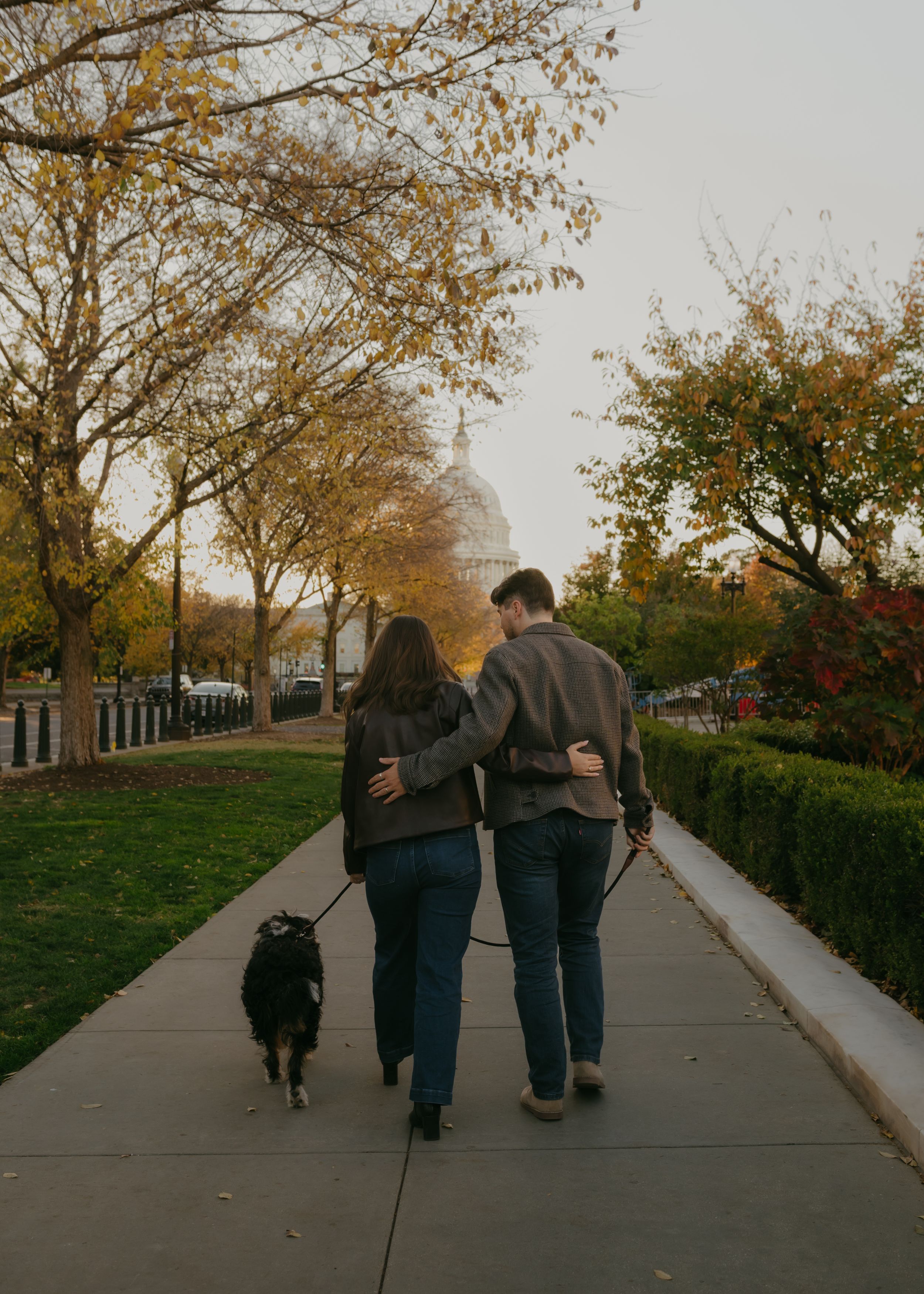 Three people and a dog walking on a concrete path surrounded by autumn trees and grass.