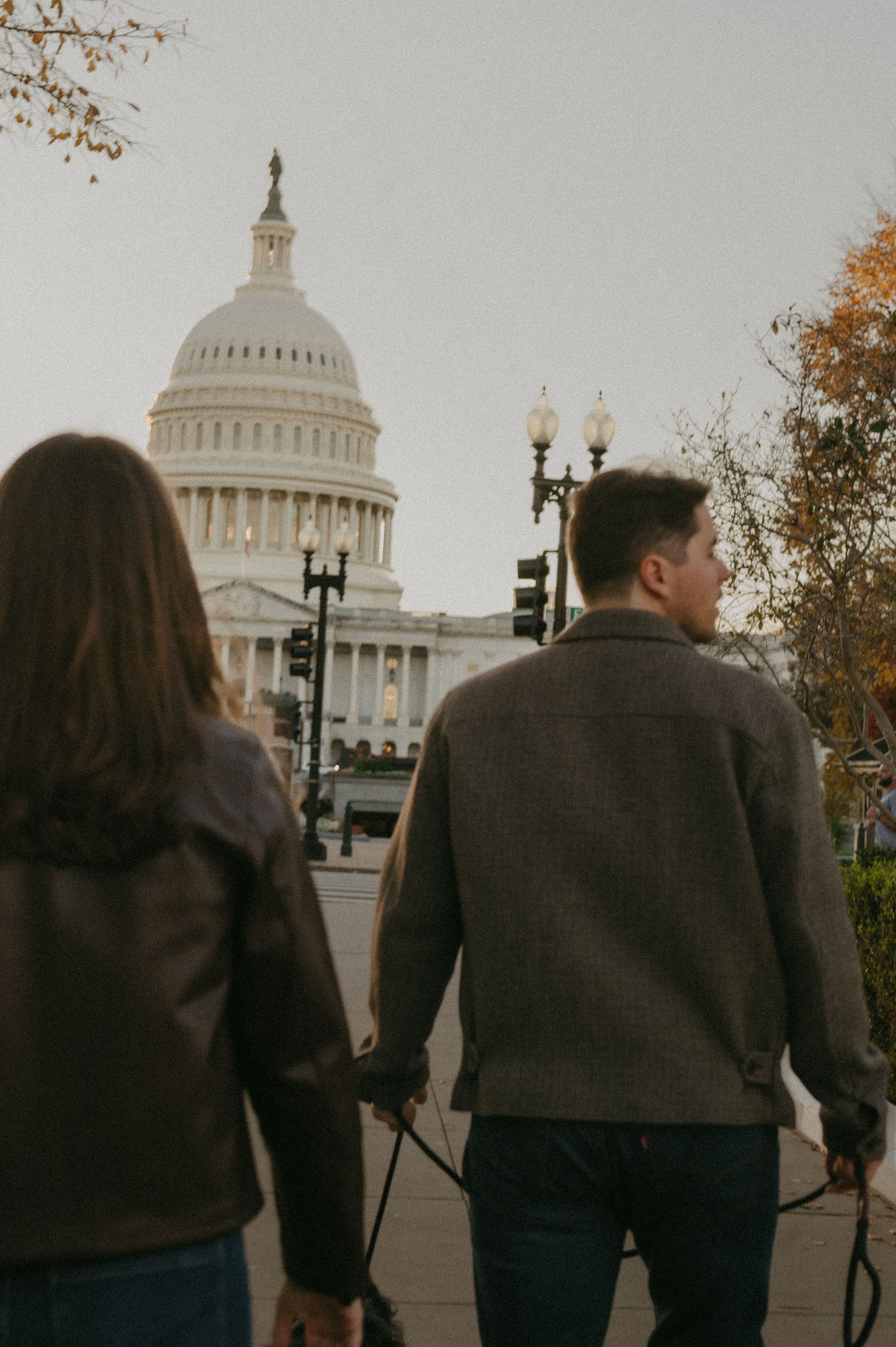 Two people in dark coats walk together towards the US Capitol building on an autumn day.