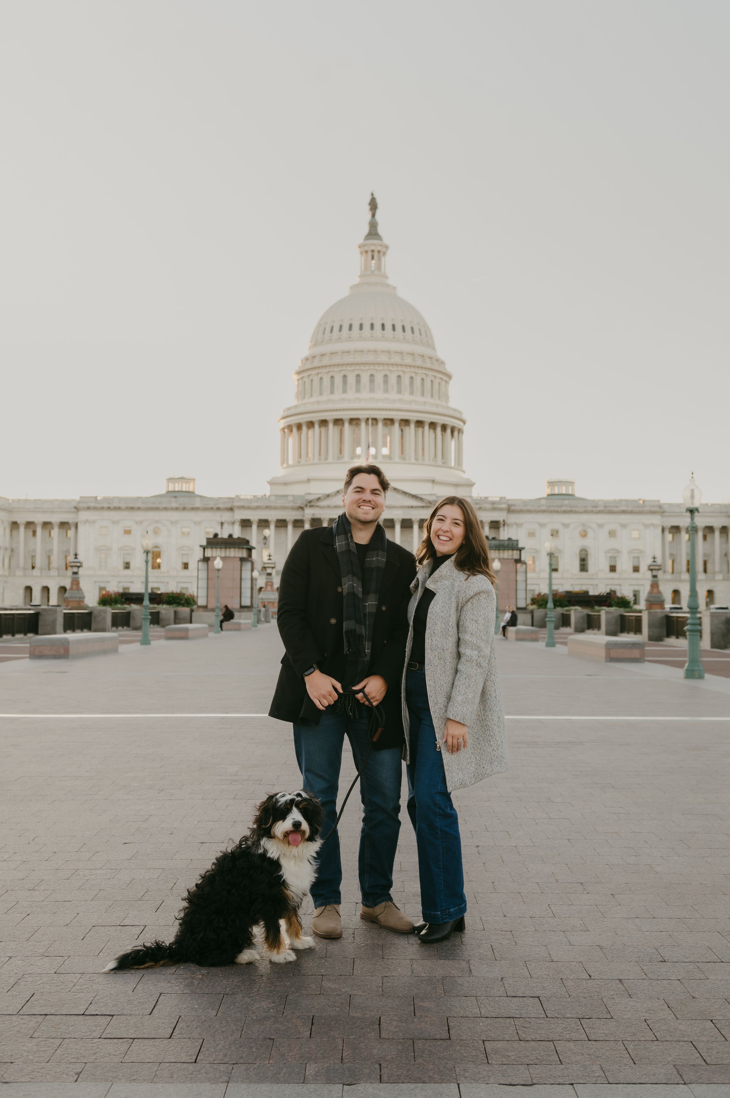 A couple and their dog pose for photos in front of the US Capitol building on an overcast day.
