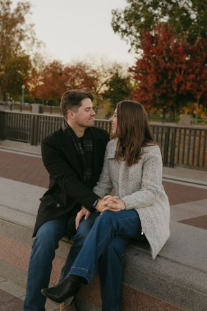 Couple sitting together on concrete ledge with fall trees and wooden fence in background.