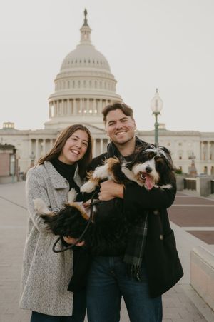 A couple holds their small dog while posing in front of the US Capitol building on an overcast day.