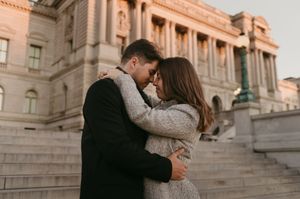 A romantic couple embraces on stone steps outside a grand historic building at sunset.