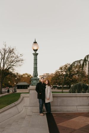 A couple stands together on a stone walkway near an ornate vintage lamp post in an autumn park setting.