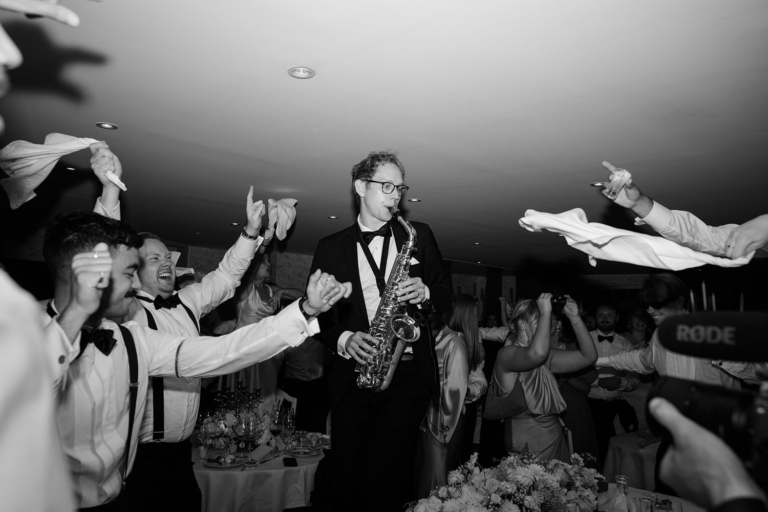 Black and white photo of a saxophonist performing at a lively party with guests raising their drinks in celebration.