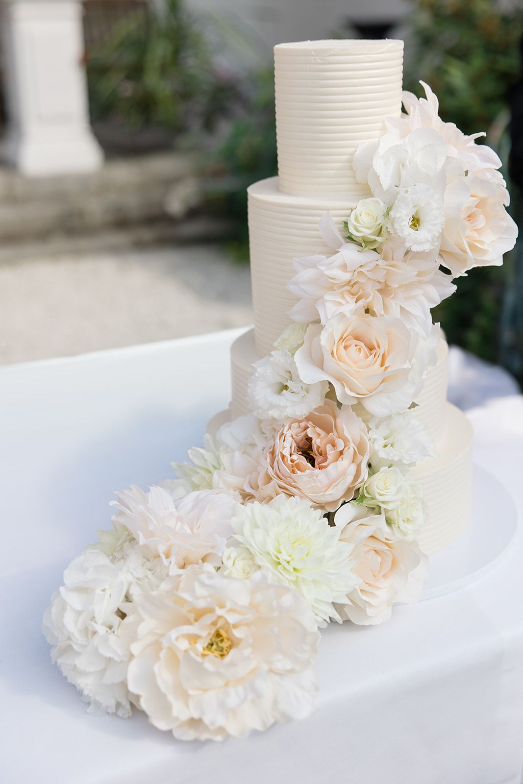 Elegant white wedding cake decorated with cascading blush roses and white flowers on a white tablecloth.