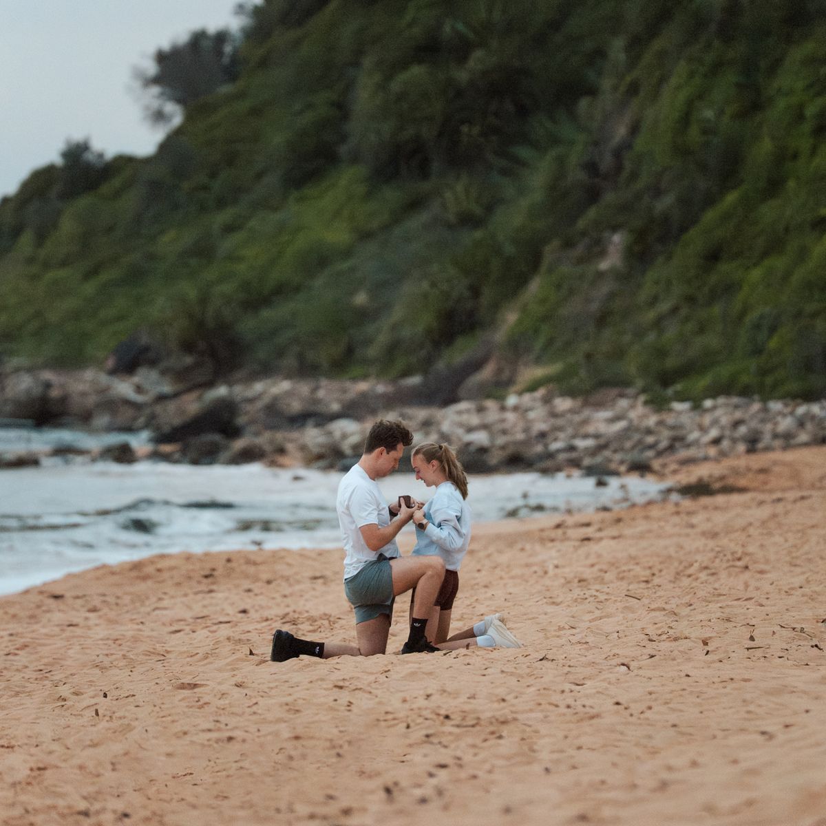 A sequence of images showing a couple kneeling on a sandy beach near rocky coastline and ocean waves during proposal photoshoot in Newport