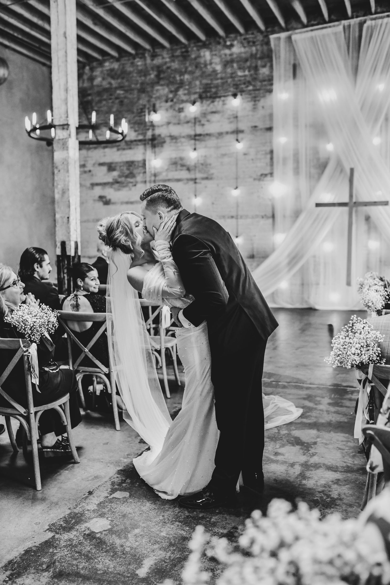 Modern editorial dip-and-kiss moment captured at Brake & Clutch Warehouse, surrounded by joyful friends and family.