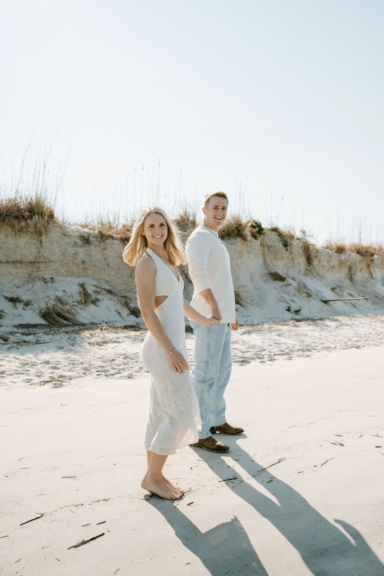 A couple walks playfully along a sunny beach with sand dunes in the background casting long shadows.