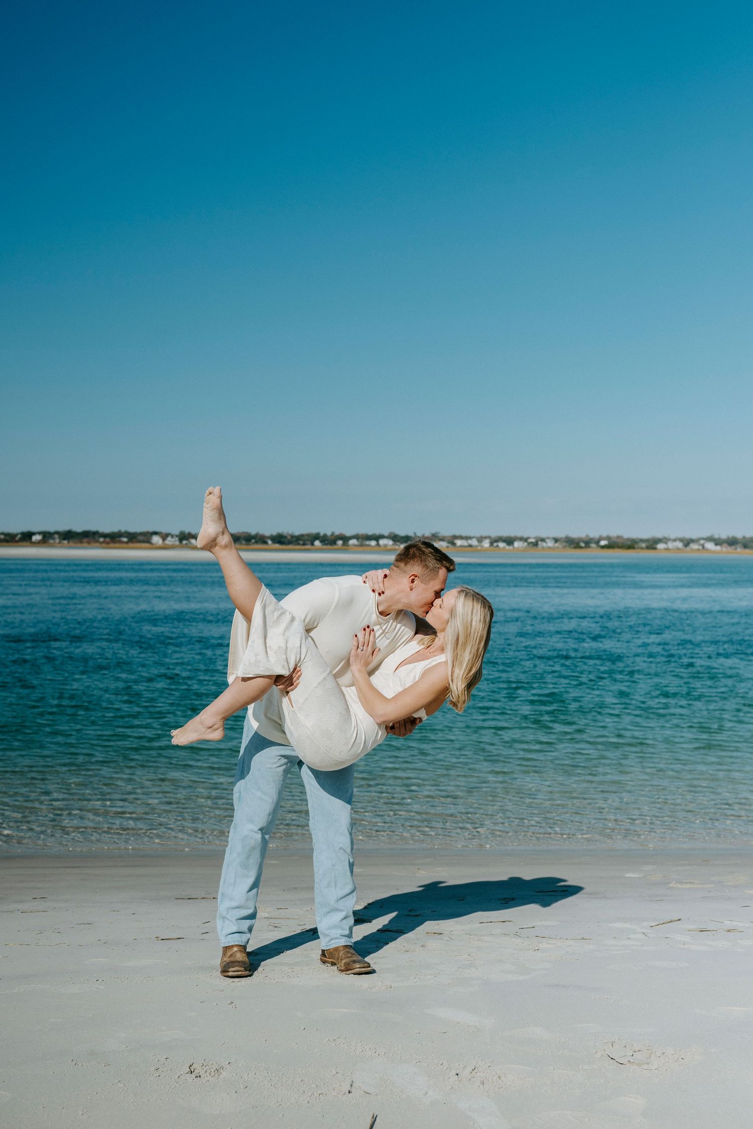 A couple embraces in a romantic dance pose on a sunny beach with turquoise waters and coastal homes in the background.