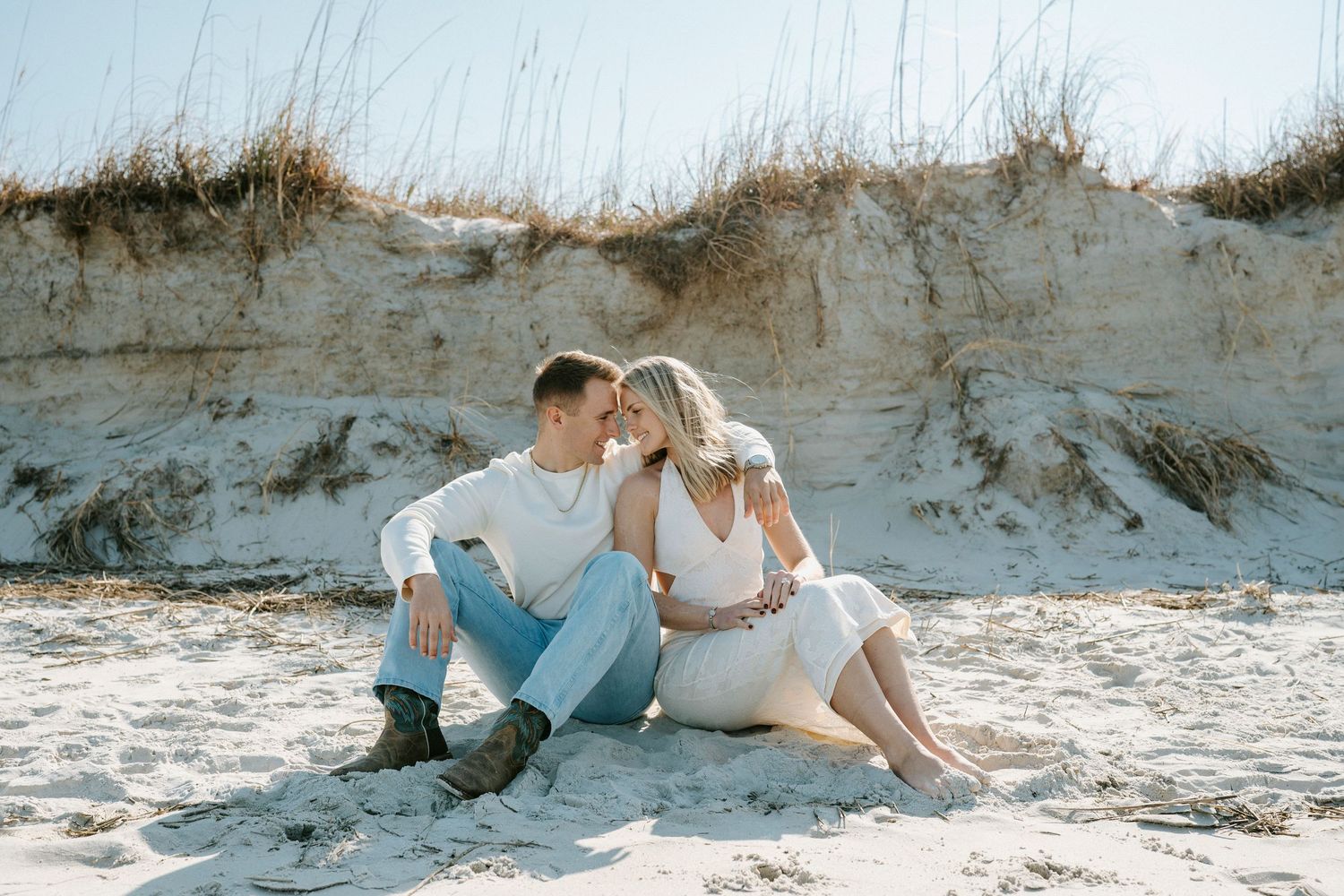 A romantic couple sits together on a sandy beach against weathered dunes on a bright sunny day.