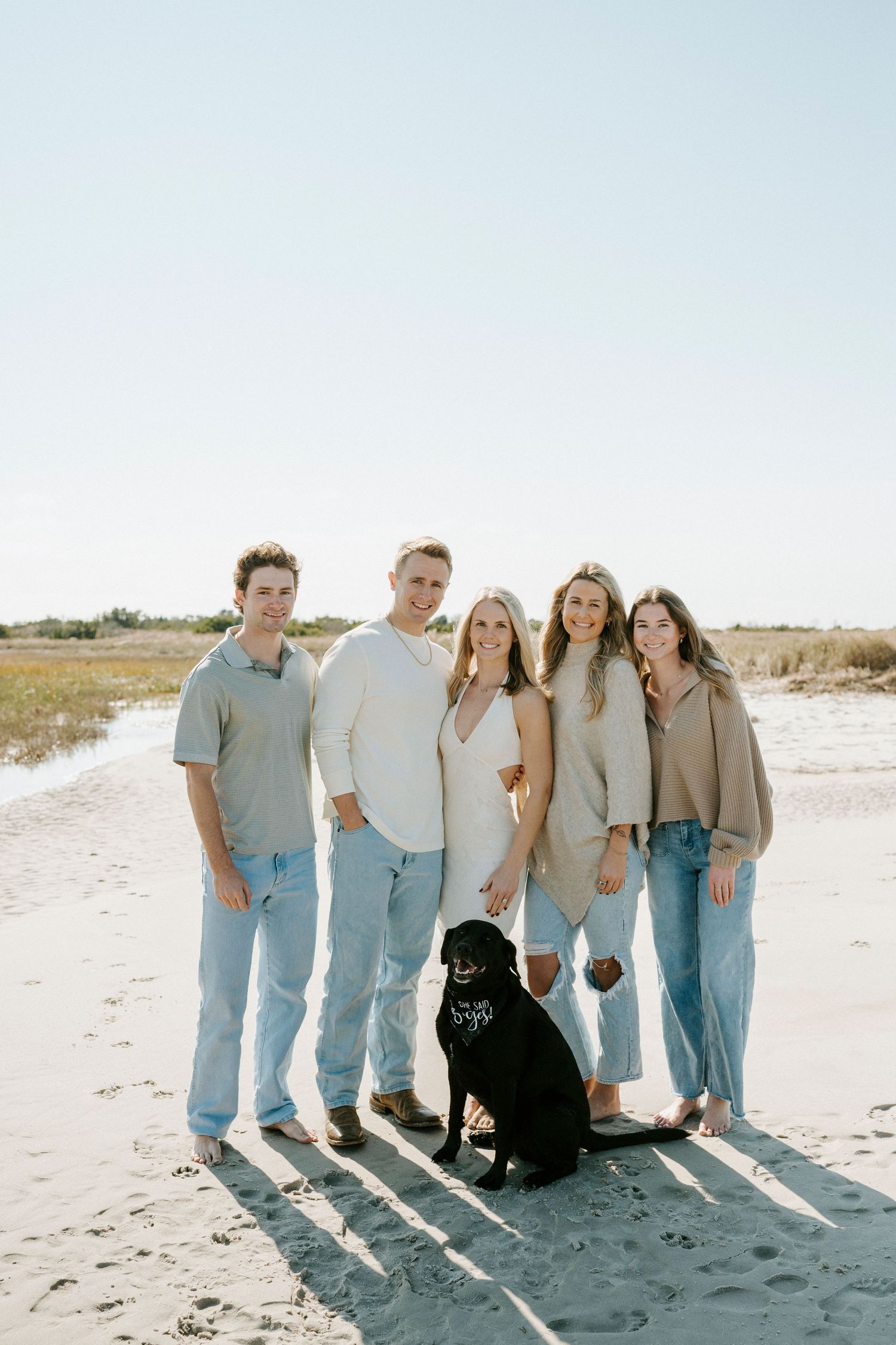 A group of people in coordinated light blue and beige outfits pose together with a black dog on a sunny beach.