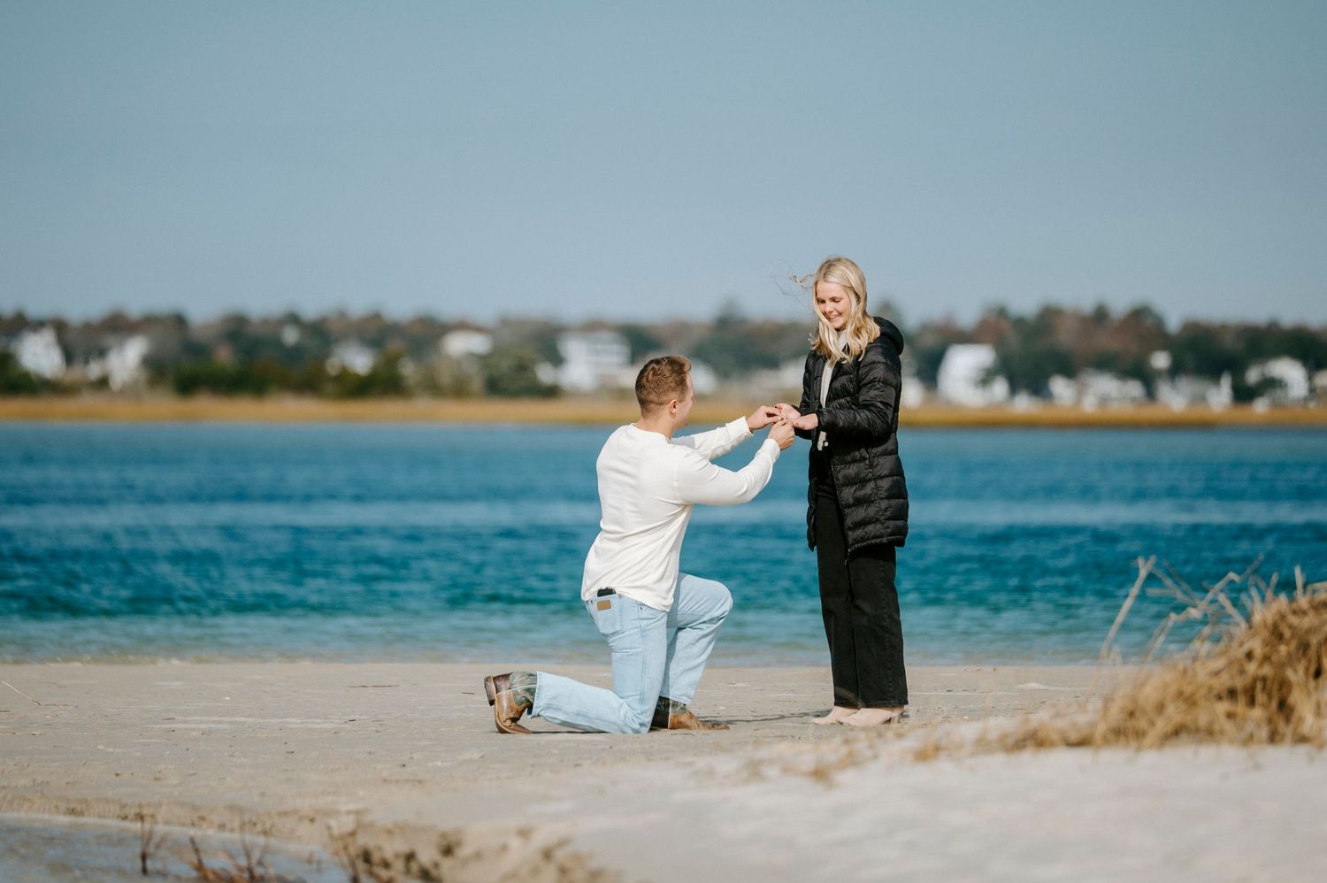 A romantic proposal scene unfolds on a beach with a scenic waterfront and residential area in the background.
