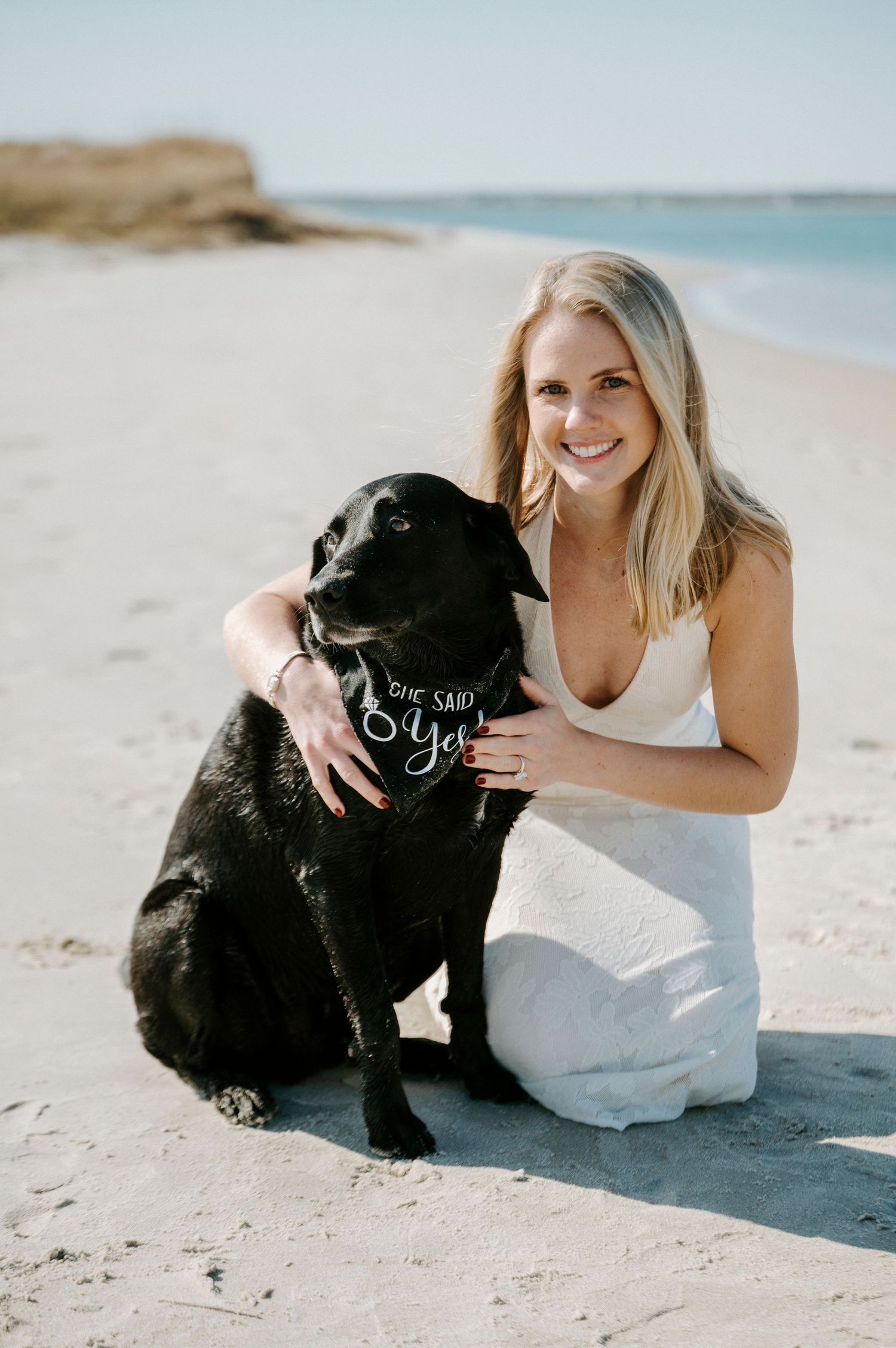 A heartwarming beach photoshoot featuring a person in white dress posing playfully with a black Labrador on sandy shores.