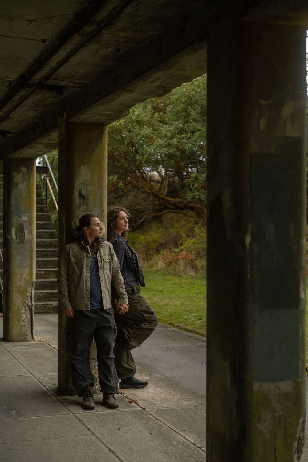 Two people sit on a bench under a concrete structure in a rural setting during overcast weather.
