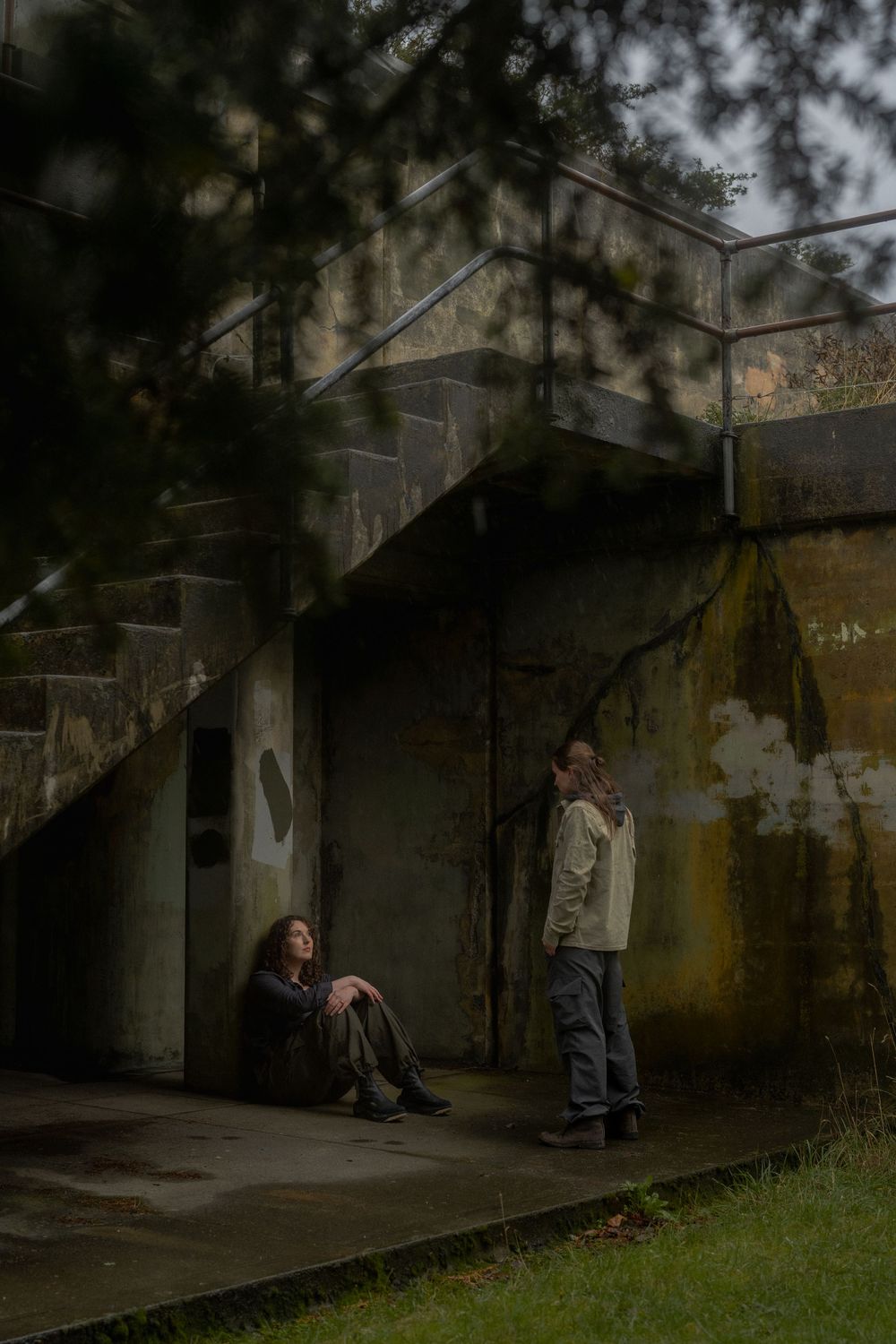 People stand in a dimly lit abandoned concrete structure with graffiti-covered walls and grass growing outside.