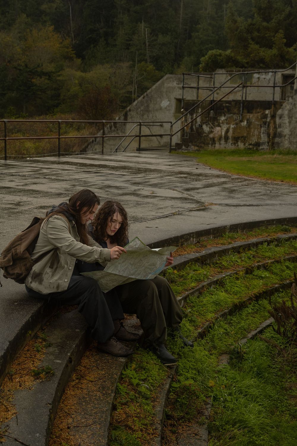 Two people sit close together on mossy concrete steps at an abandoned outdoor amphitheater on a gloomy day.
