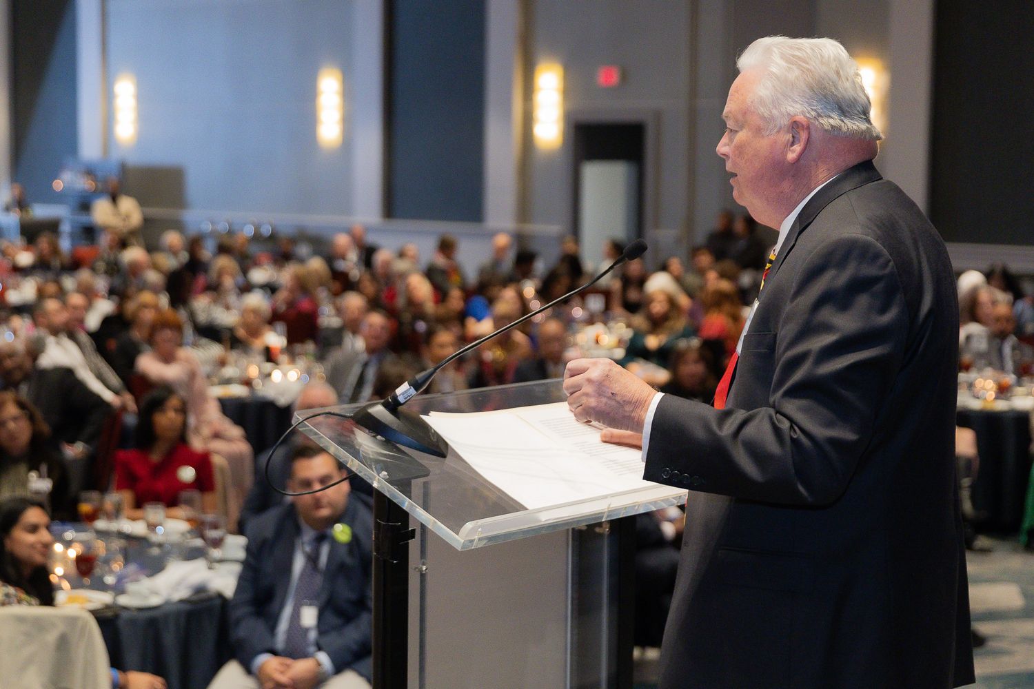 A speaker presents at a business conference to a large audience in a modern conference hall.